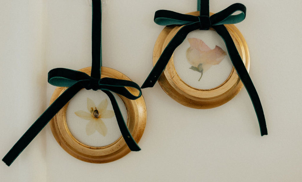Close up of two round pressed floral custom ornaments showing gold frames and dark green velvet bows on cream background