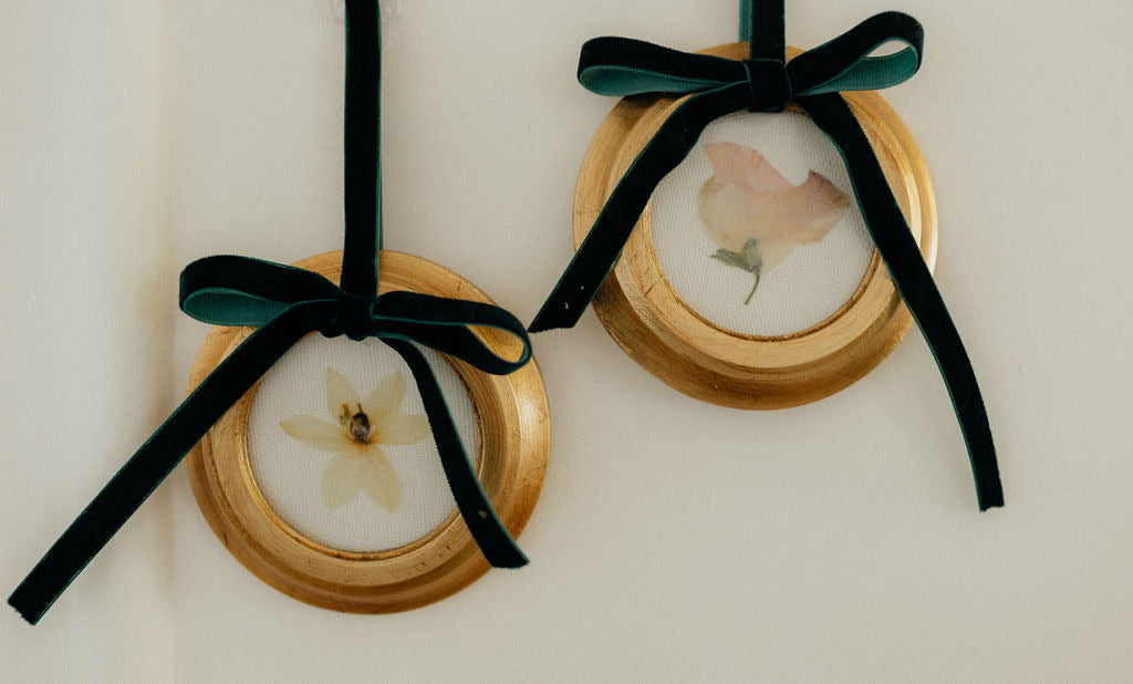 Close up of two round pressed floral custom ornaments showing gold frames and dark green velvet bows on cream background
