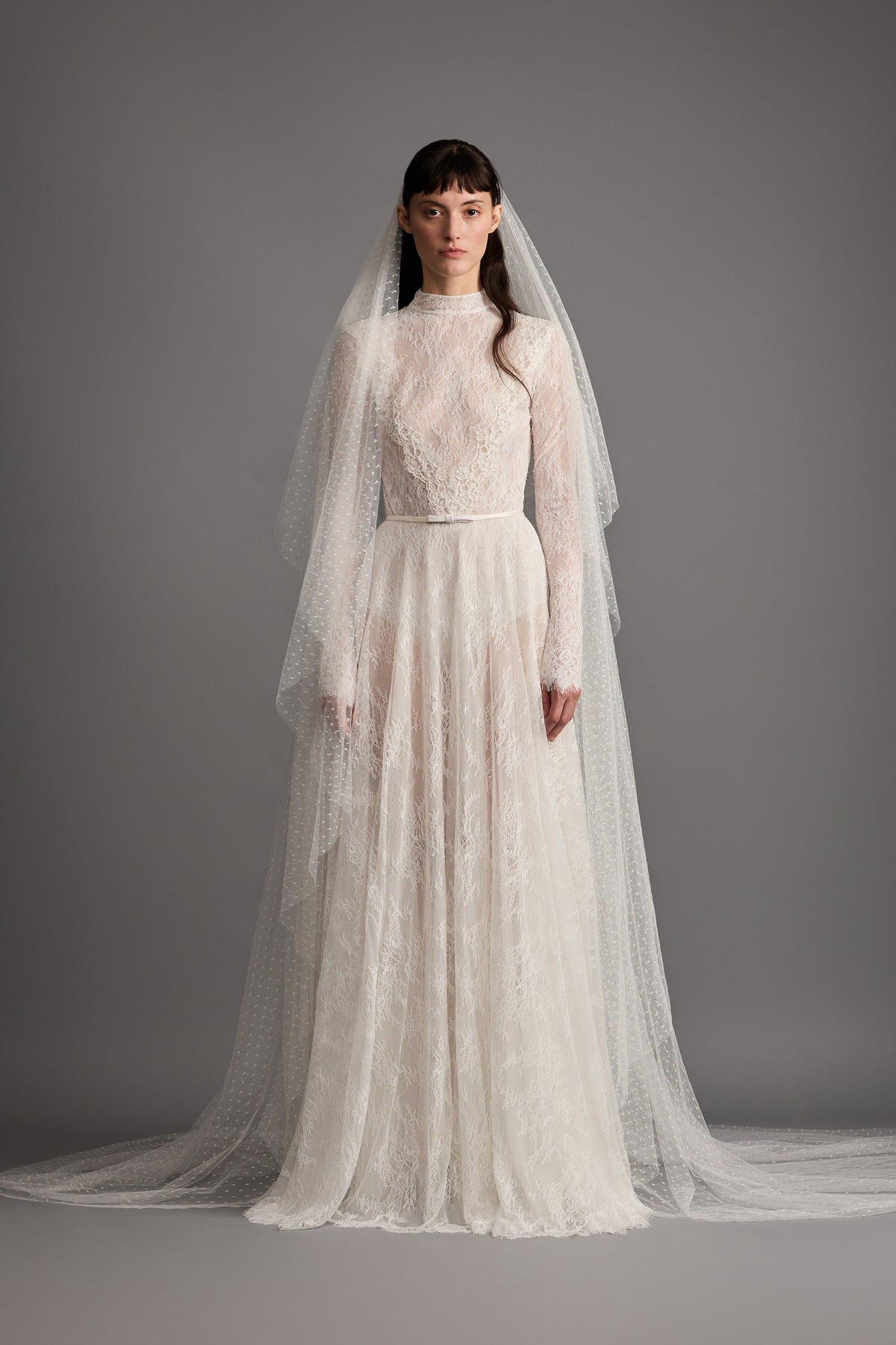 Front of Fleur Gown in warm white, A-line fit, long-sleeve lace bodice, satin bow belt and dotted tulle veil on gray backdrop