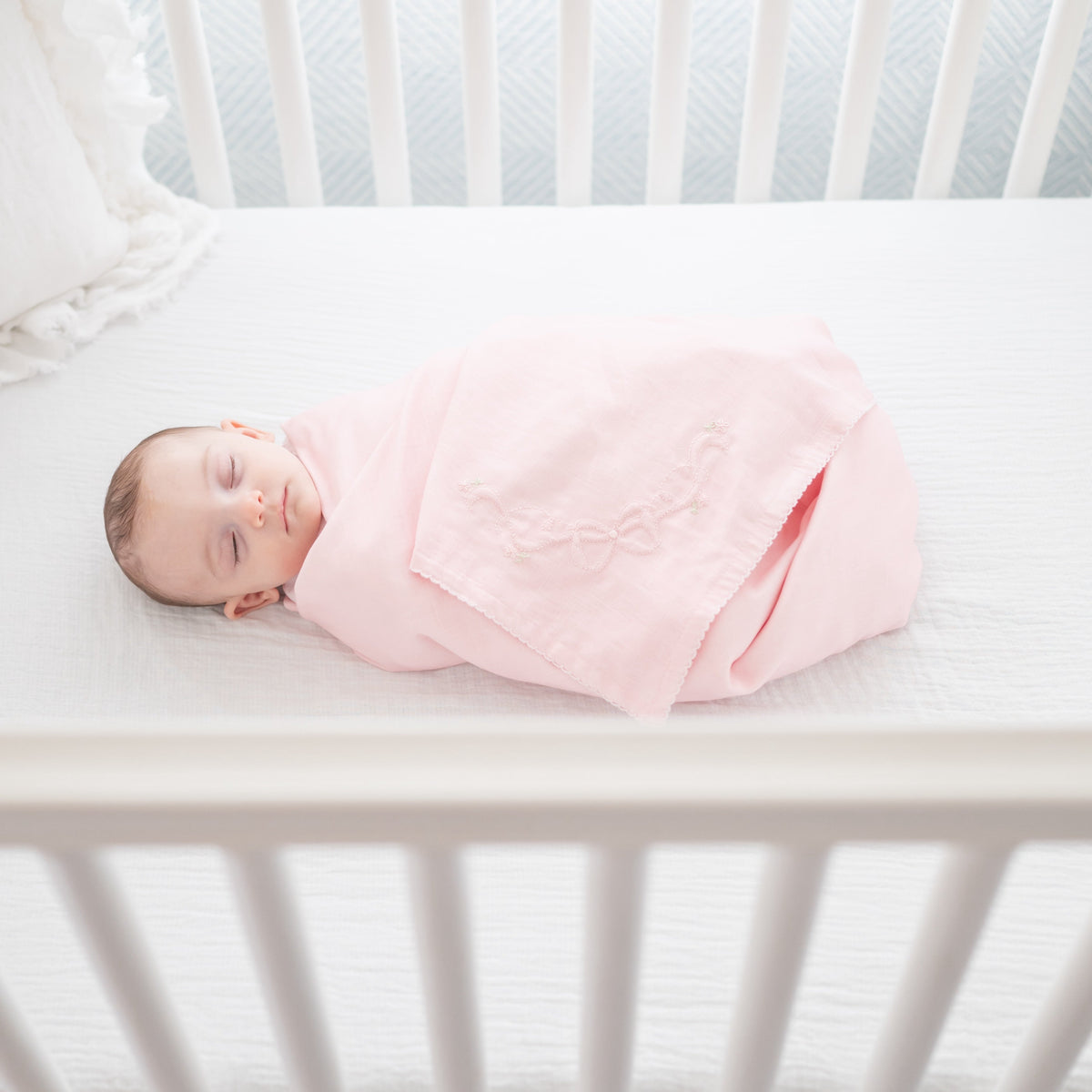 Overhead view of receiving blanket swaddling a sleeping newborn, soft pink cotton with delicate embroidered bow detail on a white crib mattress