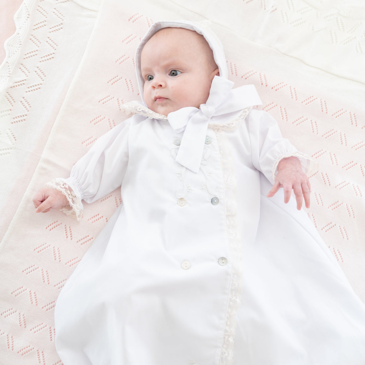 Overhead view of newborn wearing white gown and bonnet with Peter Pan collar, lace trim and buttons on pale pink blanket