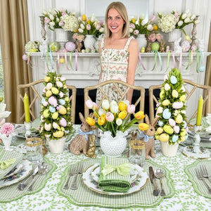 Woman standing in front of a decorated Easter table with floral arrangements and bunny decorations. Flying Sheep Country founder. Luxury table linens. Green gingham luxury embroidered placemats and napkins, green satin napkin bow ties. hand block printed green leaves tablecloth- perfect for luxury easter entertaining. 