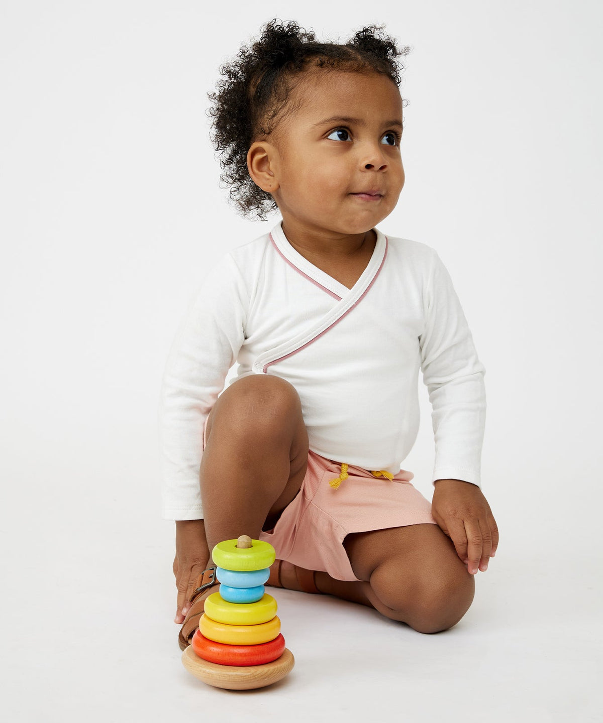 Front three-quarter view of Crossbody One-Piece in white with rose piping, long sleeves, worn by a toddler kneeling on a white background