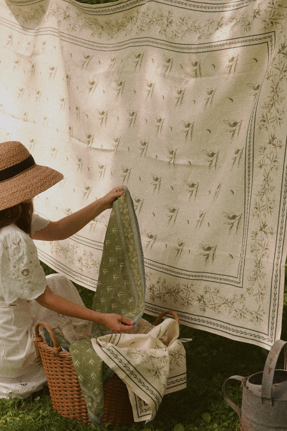 Woman holding a green olive branch printed linen hand towel beside a hanging matching linen in a grassy garden