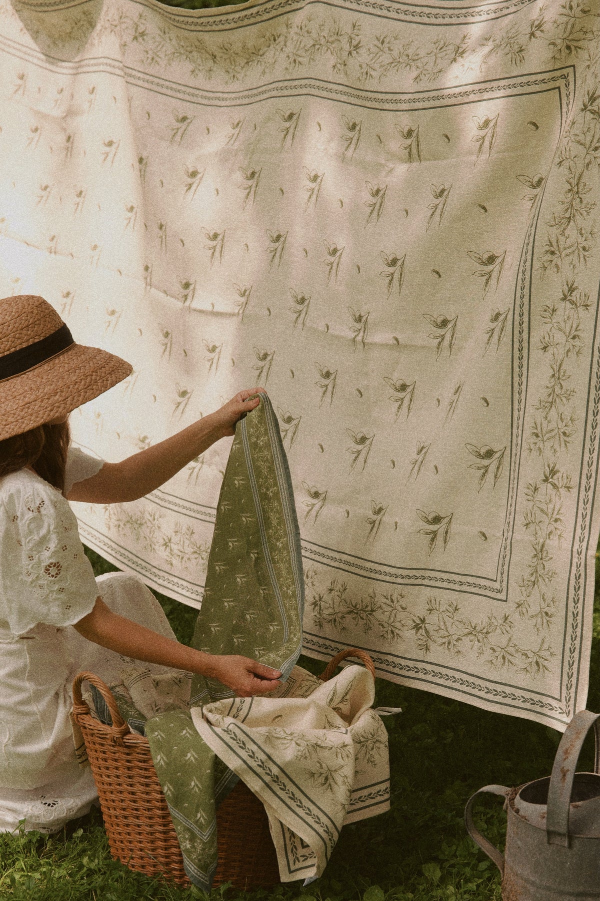Woman holding a green olive branch printed linen hand towel beside a hanging matching linen in a grassy garden