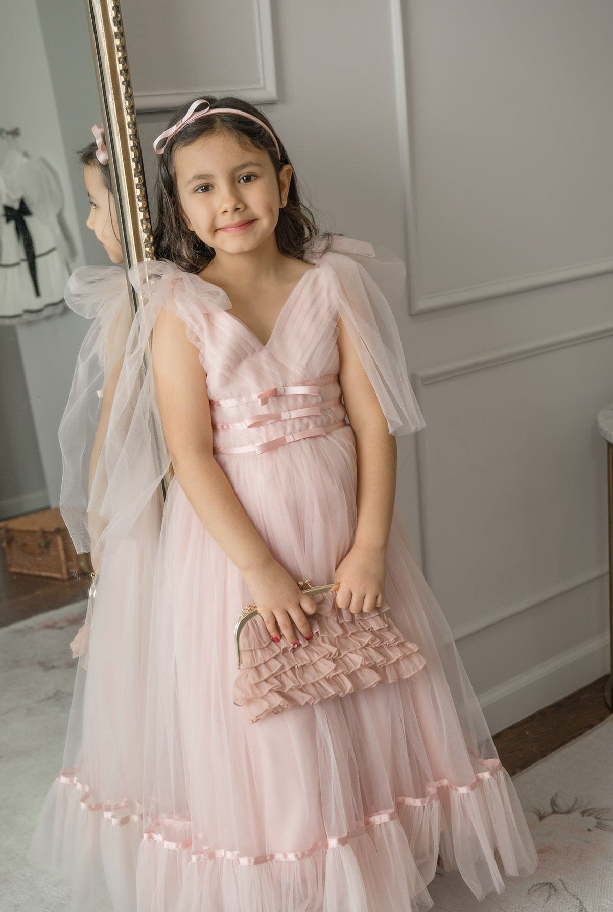 Light pink tulle flower girl dress with V-neck bodice and soft tiered skirt, styled indoors by mirror.