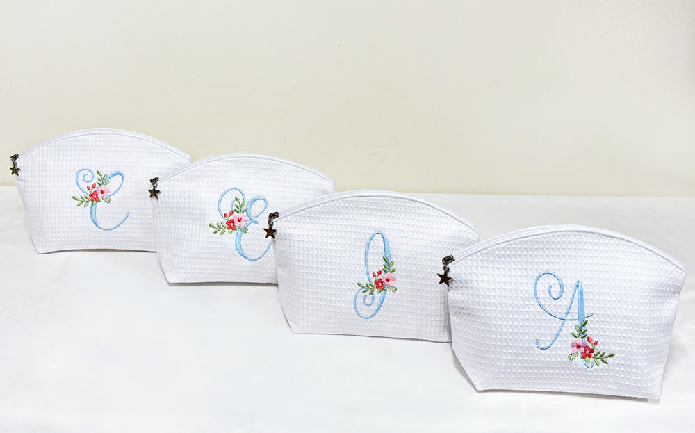 Front view of four white waffle-weave cosmetic bags with blue embroidered monograms, floral accents, and star zipper pulls