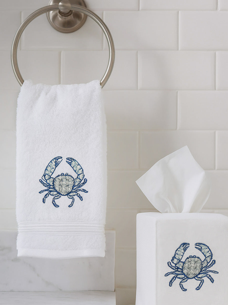 Front view of guest towel with blue embroidered crab on white terry, hanging on a metal towel ring in tiled bathroom