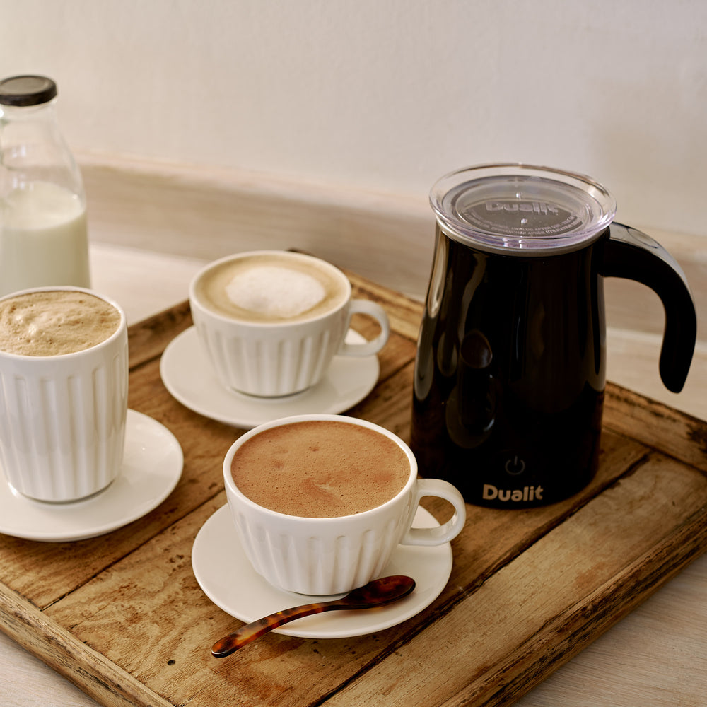 Three-quarter view of black milk frother and hot chocolate maker with clear lid and handle on wooden tray beside three cups