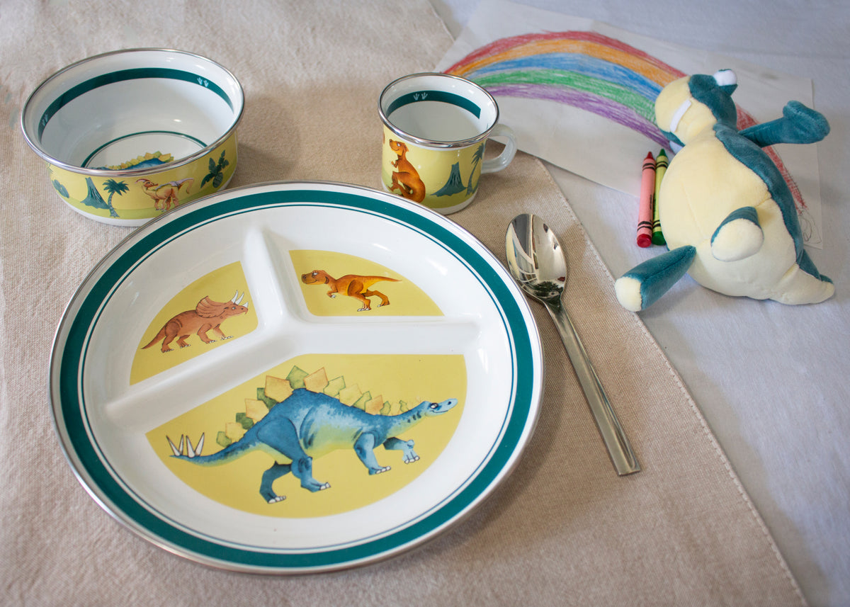 Angled view of Child Set in Dinosaurs with dinosaur illustrations on a mug, bowl, and divided plate on a cloth-covered table
