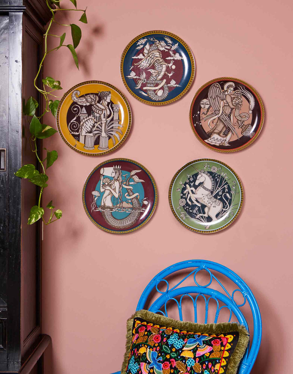 Five decorative plates, including the light green unicorn design, on a pink wall above a blue chair with an embroidered pillow