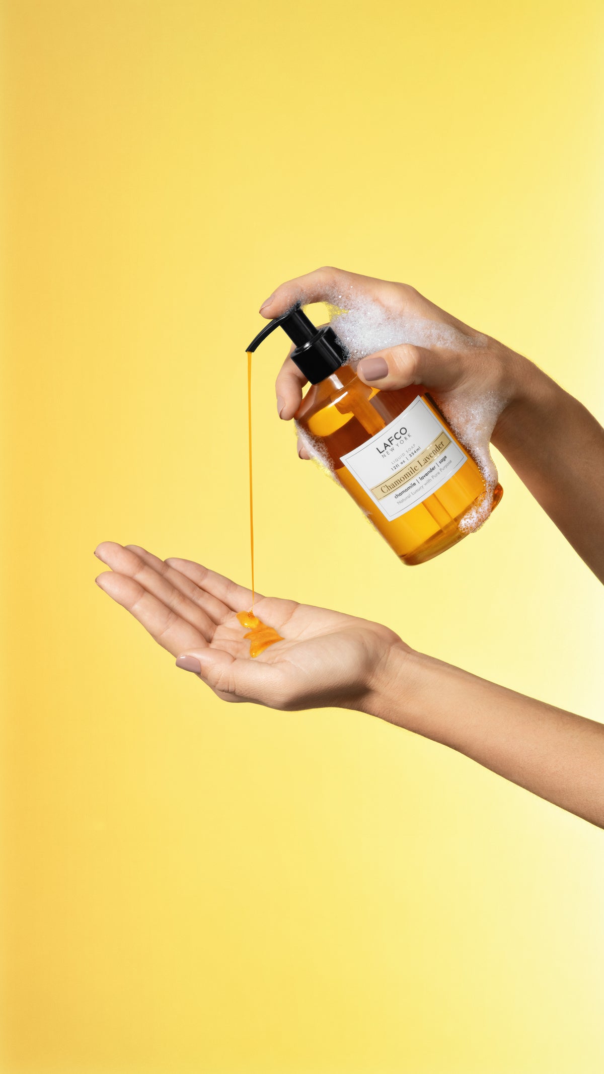 Hand holding Chamomile Lavender Liquid Soap bottle dispensing amber soap into cupped hand with foam on yellow background