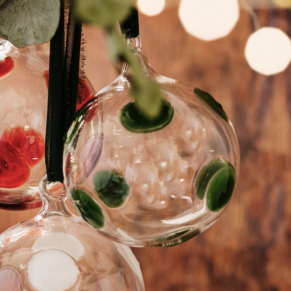 Close-up of Drop ornament in green showing clear mouthblown glass with raised green dots and black ribbon against a warm blurred wood background