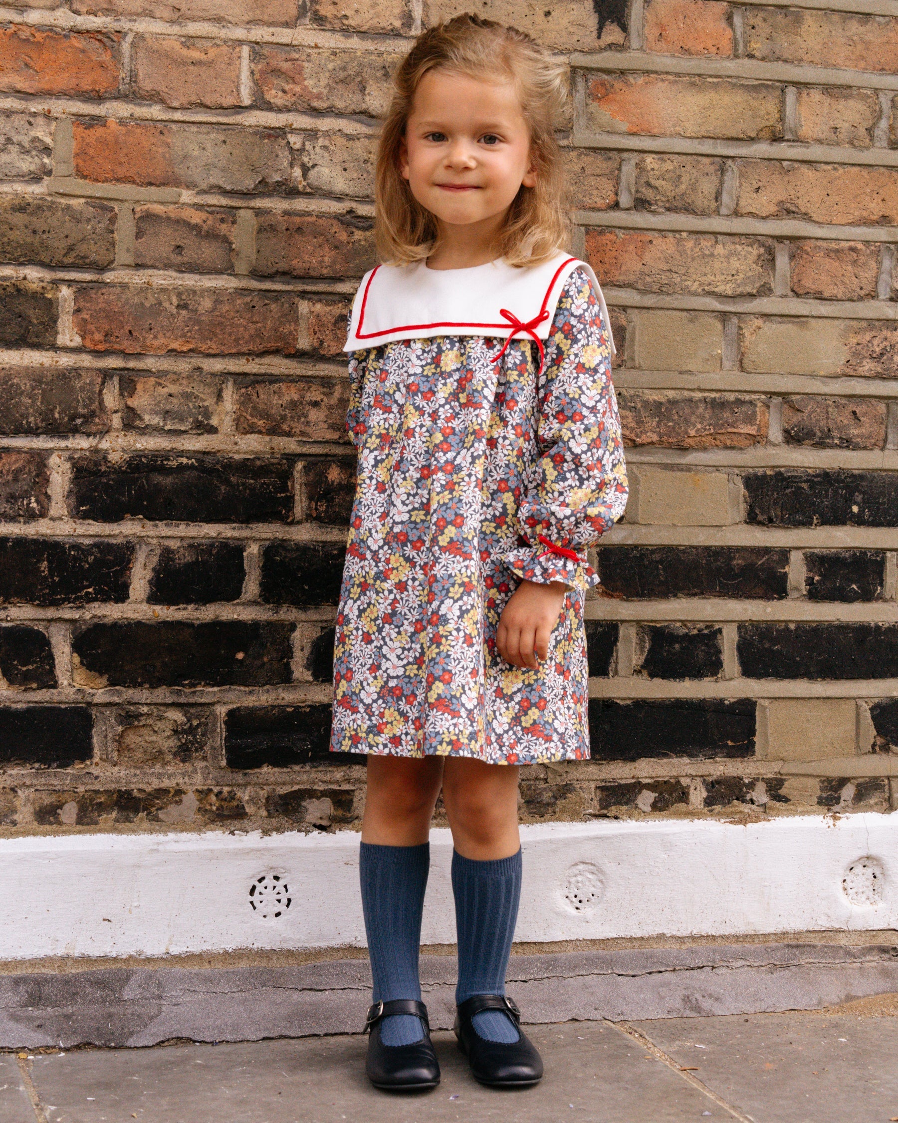 Young girl in a floral dress with a red collar standing against a brick wall.