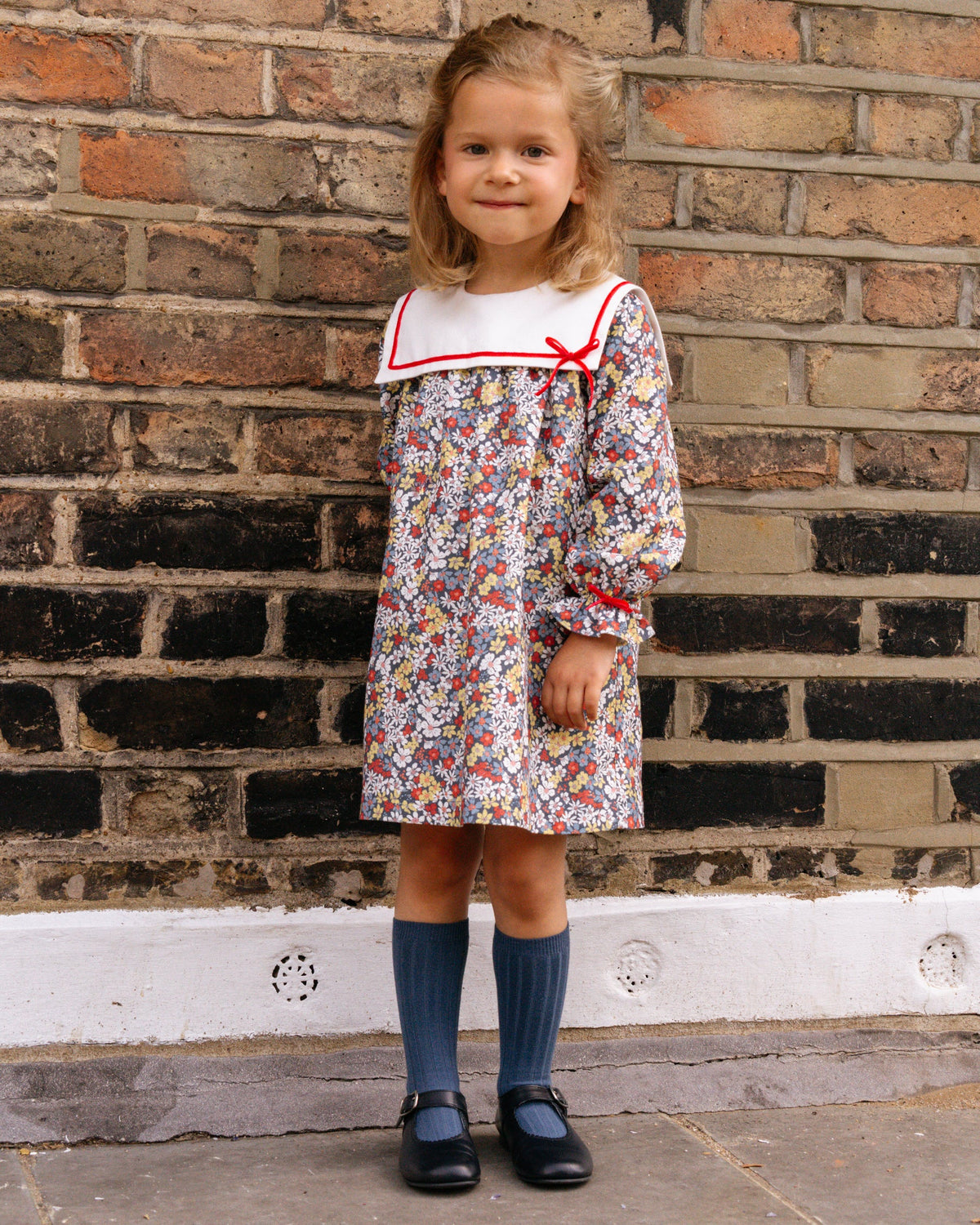 Young girl in a floral dress with a red collar standing against a brick wall.