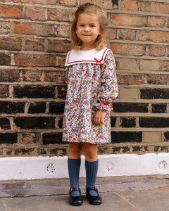 Young girl in a floral dress with a red collar standing against a brick wall.