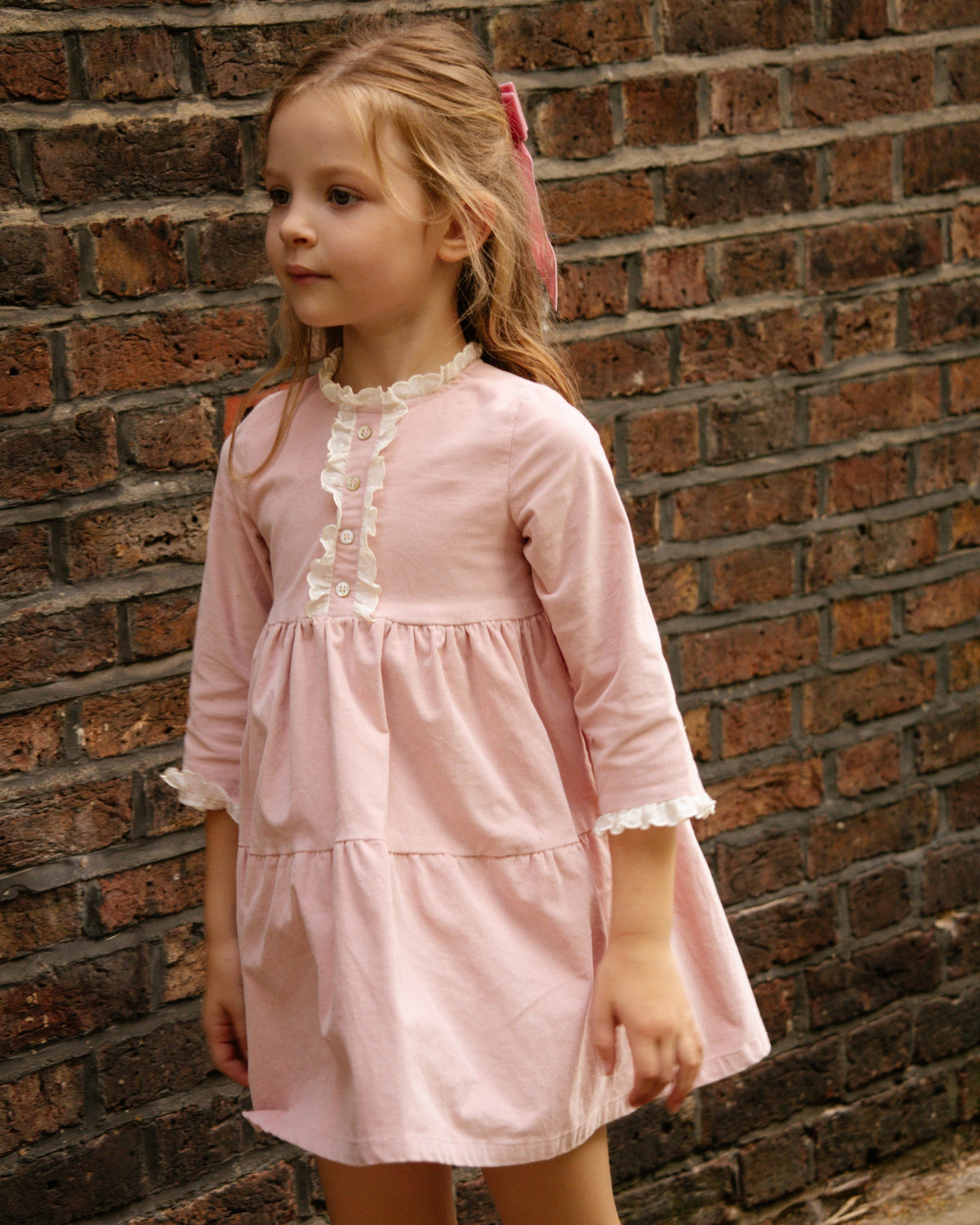 Young girl in a pink dress standing against a brick wall