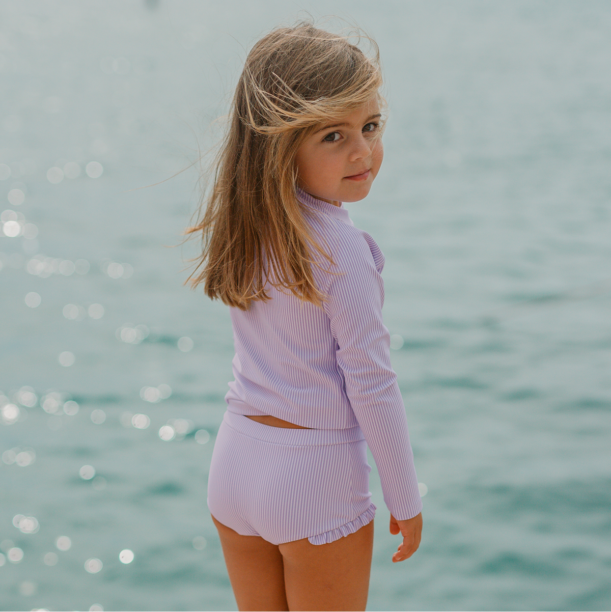 Back view of Girls Lavender Stripe Rashguard Set showcasing long sleeves and ruffle hem with ocean background