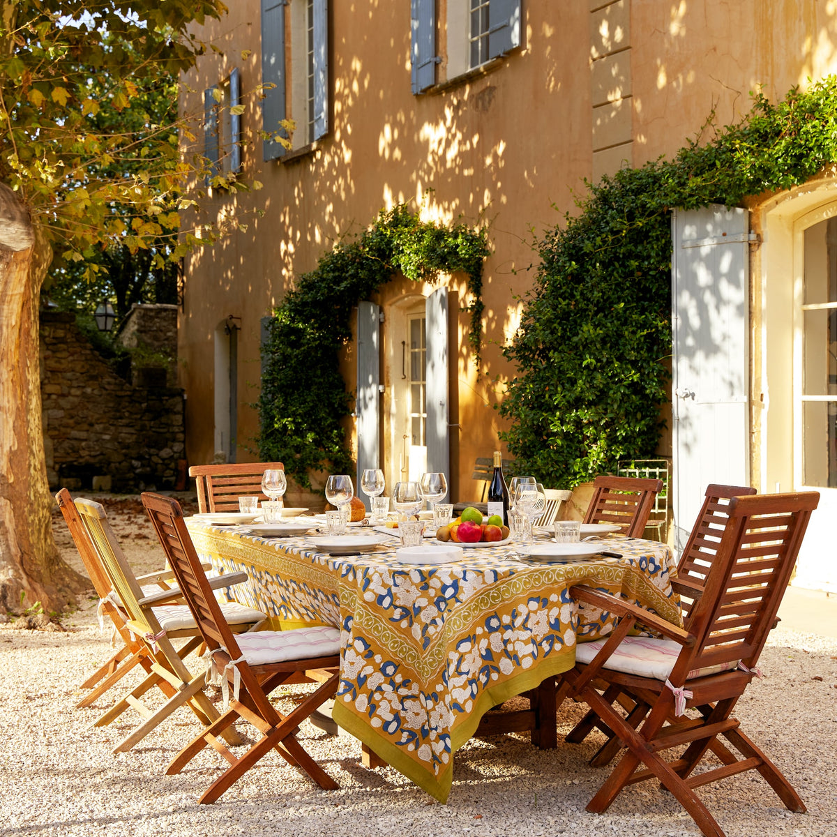 Outdoor dining table set with the Forest Harvest mustard and blue tablecloth in a sunlit Provençal courtyard