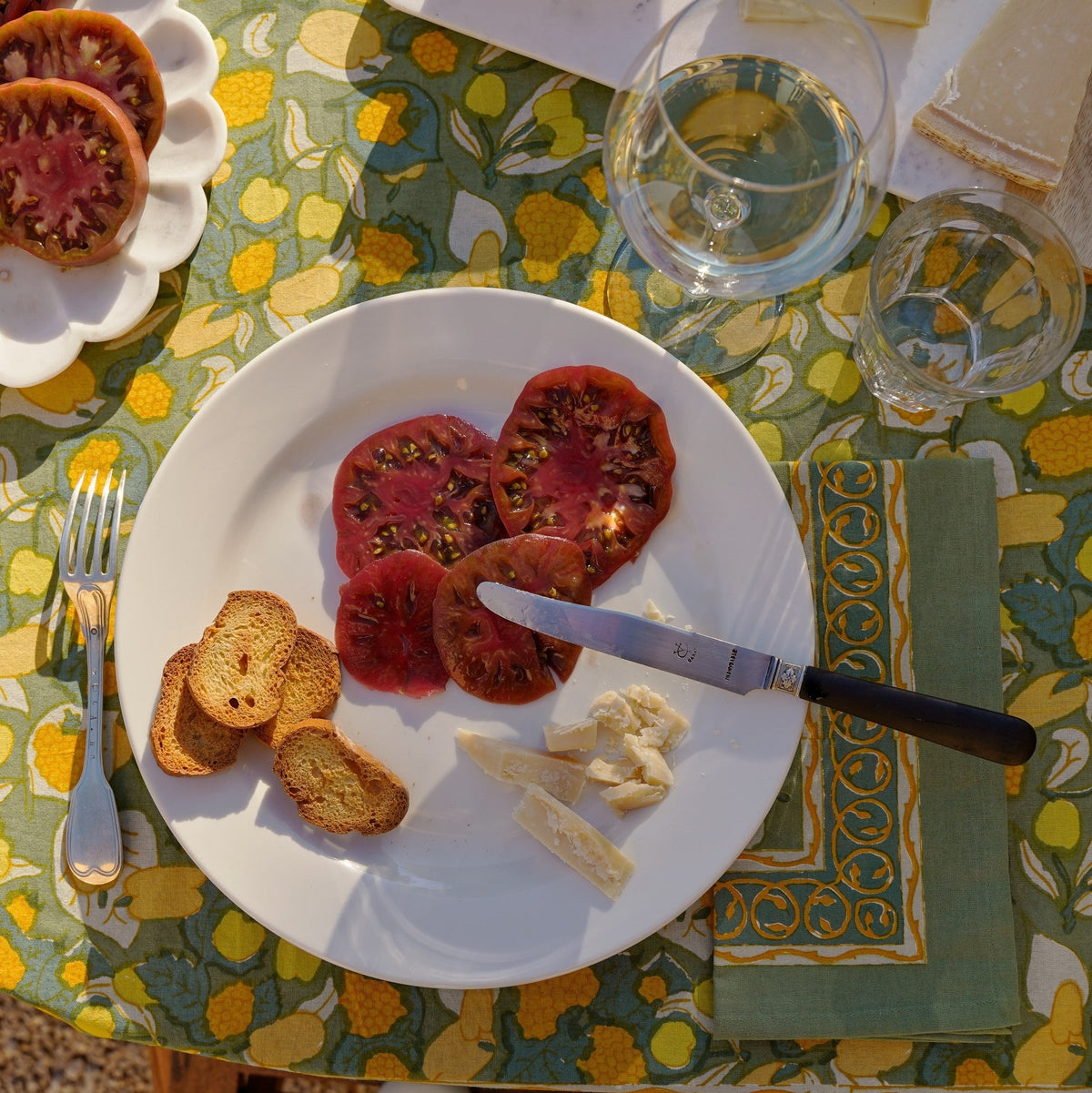 Fruit-print yellow and green linen napkin beside a white plate of sliced tomatoes, cheese, and toasted bread on a sunlit outdoor table