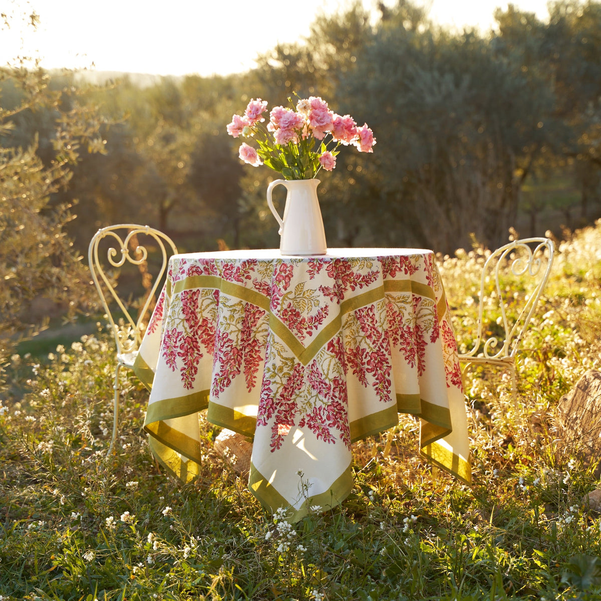 Front view of French Tablecloth Wisteria Green and Pink showcasing olive green vines and pink flowers with a garden background