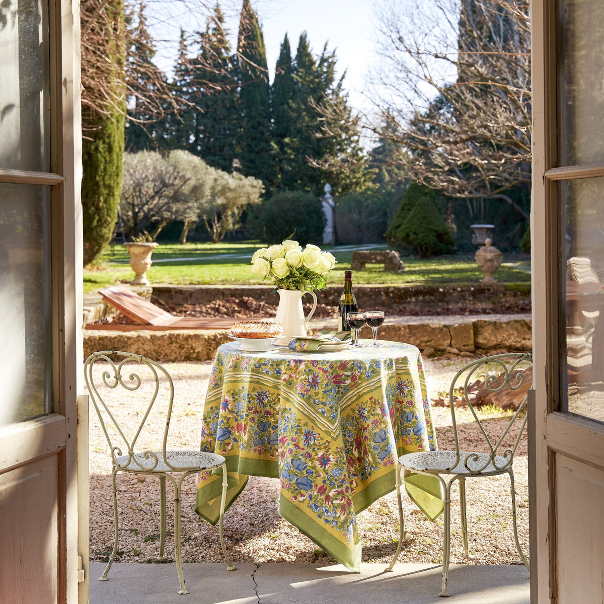 French Tablecloth Jardin In Blue and Vert covers a dining table with flowers and wine in a sunny garden setting