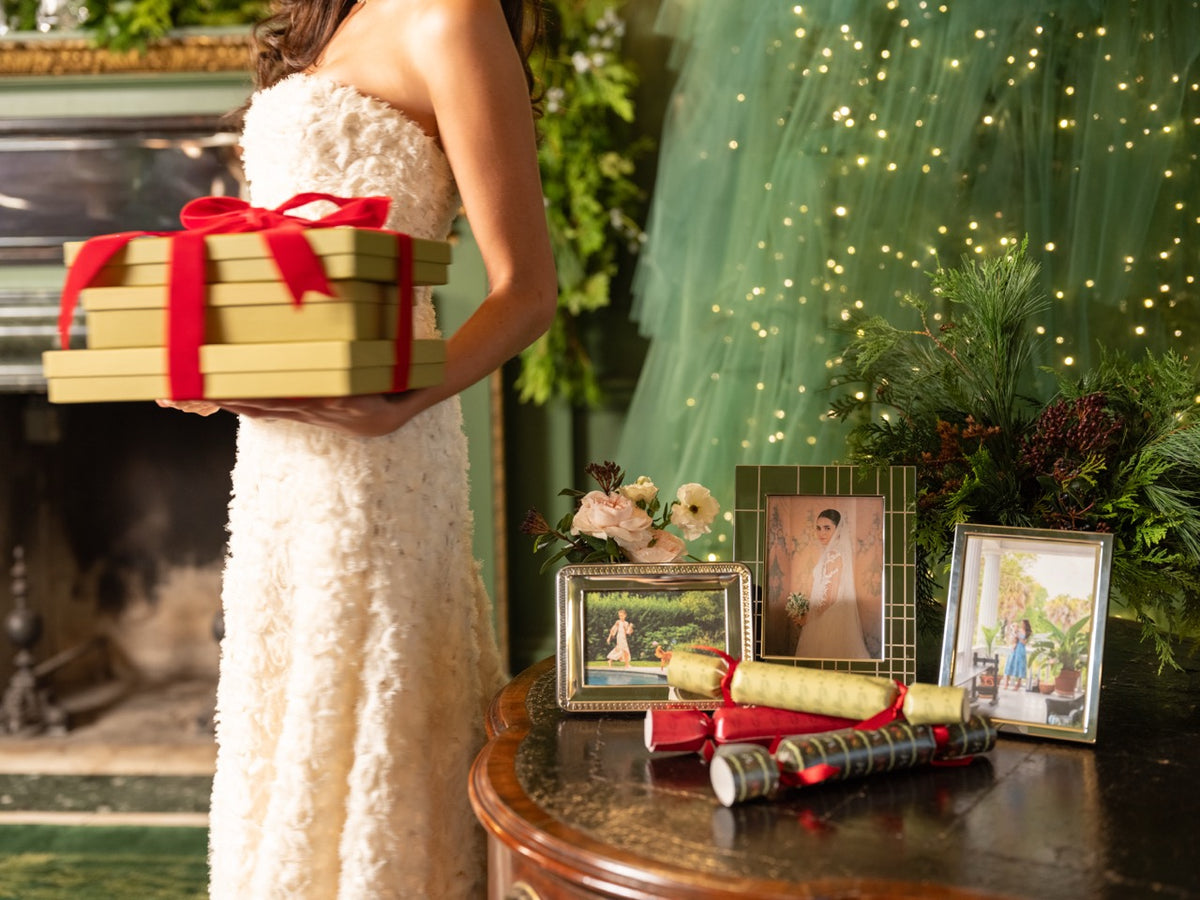 Woman in a white dress holding wrapped gifts beside a table displaying picture frames and holiday decorations