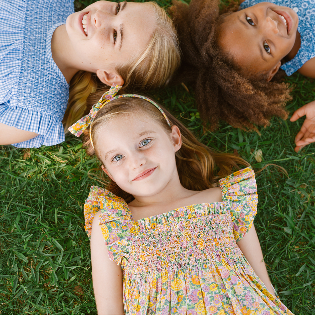 Three girls lying on grass, with one wearing the Liberty fabric headband from the Girls Headband and Scrunchie Pack