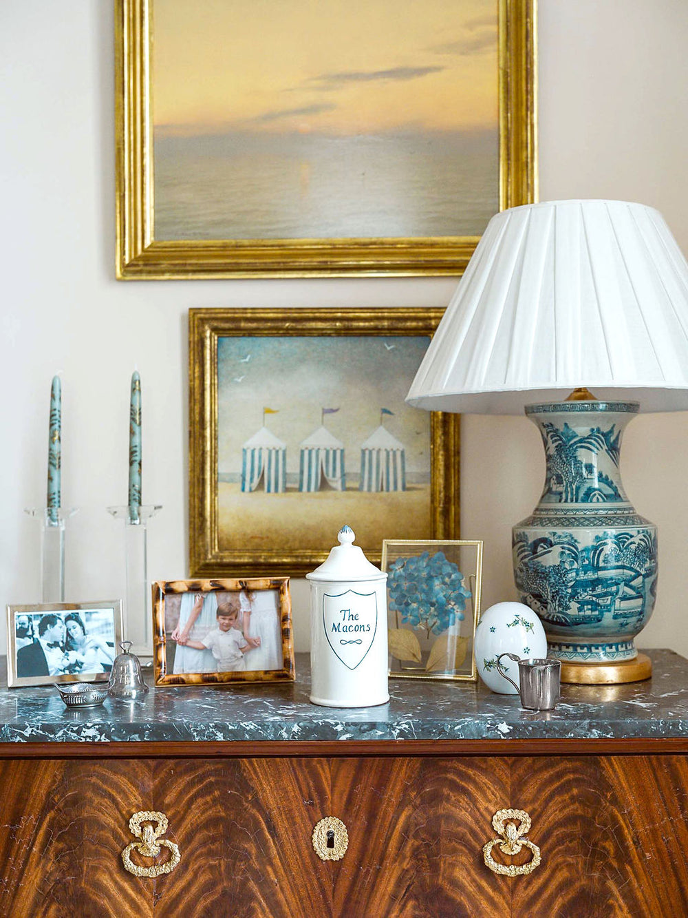 White hand-painted apothecary jar monogrammed The Macons on a marble dresser with framed photos and a blue porcelain lamp