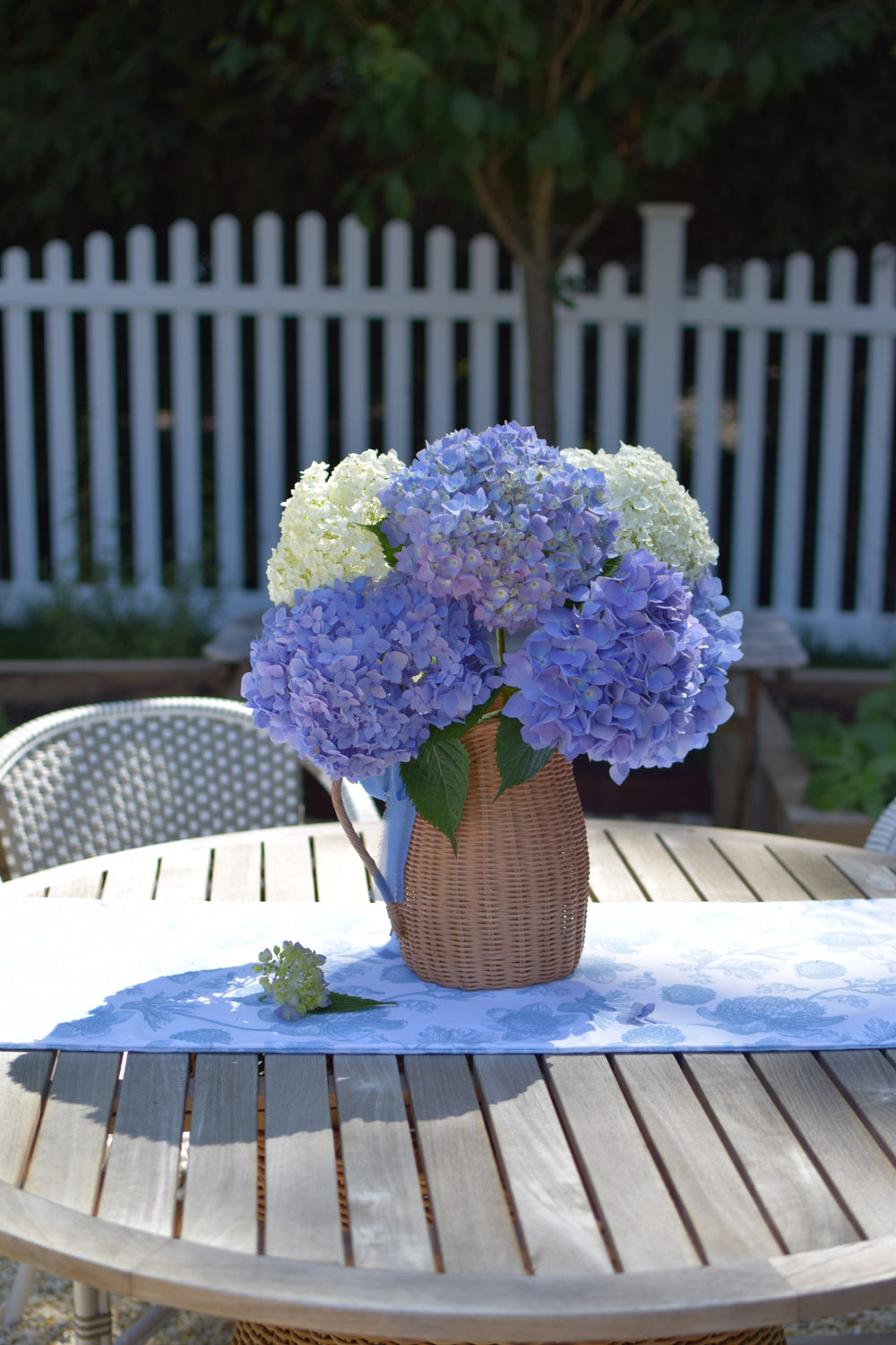 Front view of Garden Toile table runner showing cornflower-blue toile print on a round wooden patio table with hydrangeas