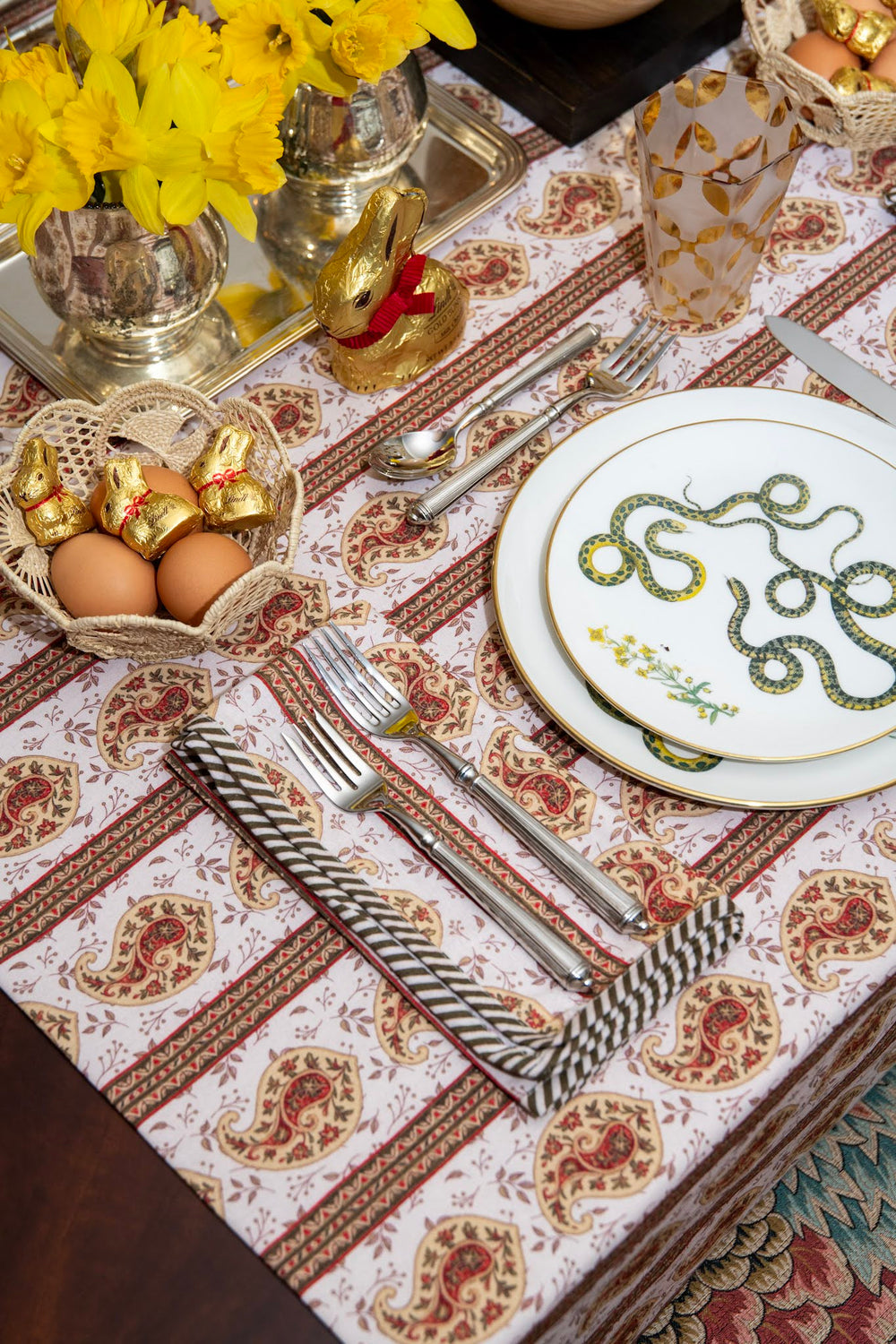 Angled view of Celina tablecloth showing olive and maroon paisley on cream with place setting and yellow daffodils