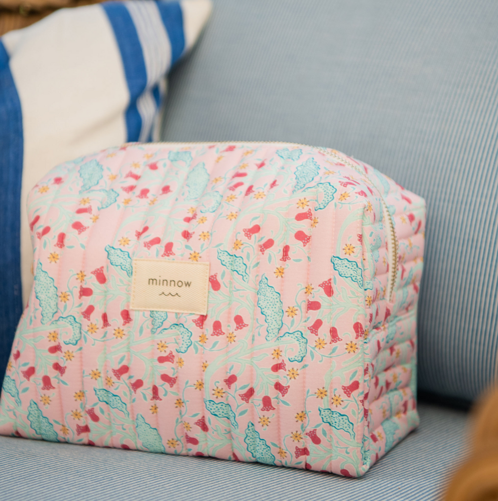 Angled front view of Bluebells travel pouch showcasing quilted pink floral print, zipper top, and minnow label on a blue-striped couch