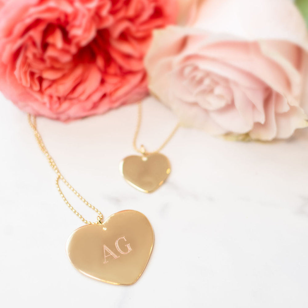 Close-up of large heart necklace showing engraved monogram AG on polished gold pendant with pink roses in the background