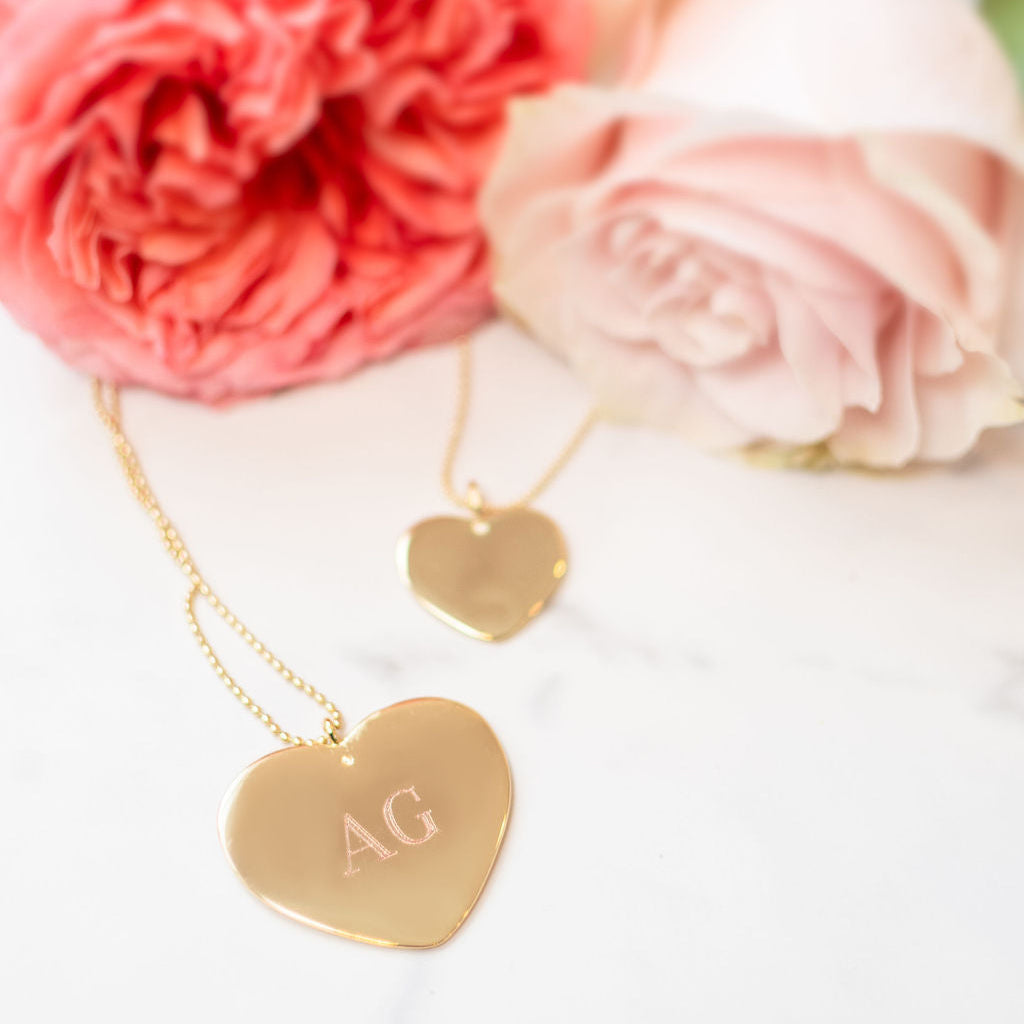 Close-up of large heart necklace showing engraved monogram AG on polished gold pendant with pink roses in the background