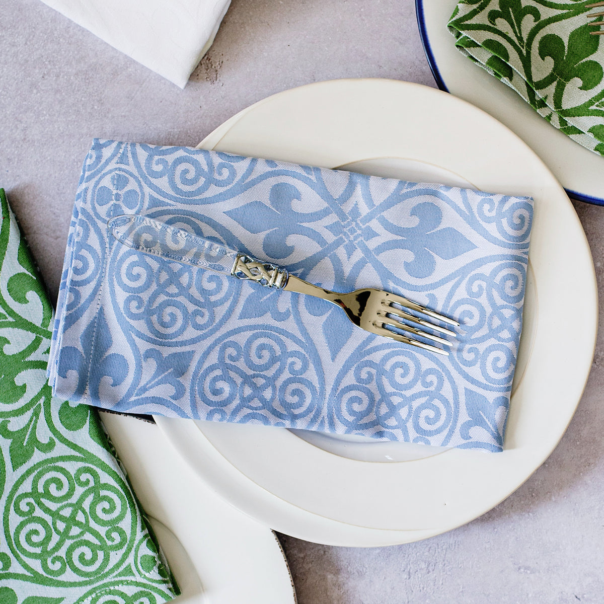 Top-down view of blue damask cotton napkin folded on white plate showing woven damask pattern with silver fork on gray tabletop