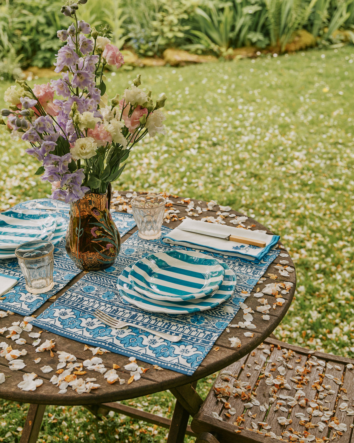D’Ascoli Malabar placemat styled on a wooden outdoor table with blue indigo floral pattern, striped plates, glassware, and a vase of pink and purple flowers