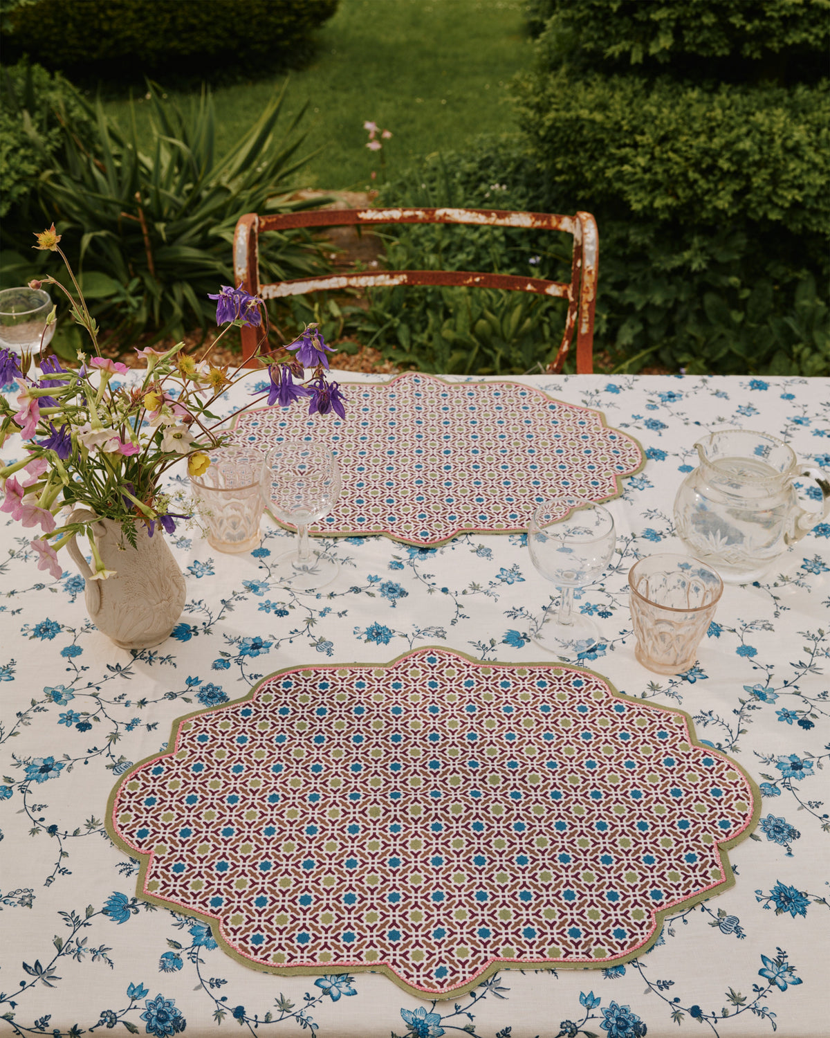 Overhead of D’Ascoli Adoria scalloped linen placemat showcasing green-trimmed geometric blue and red embroidery on a blue floral tablecloth in a garden setting