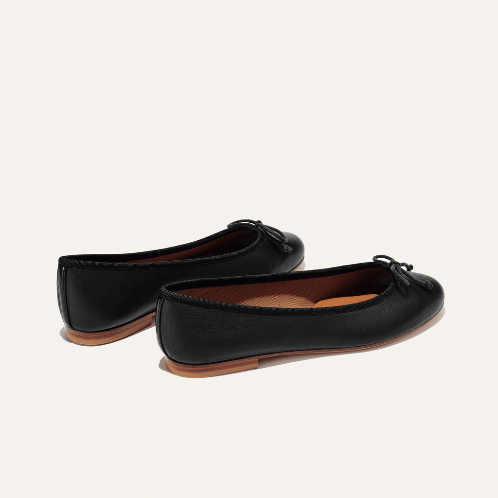 The Demi - Black Nappa by margaux-development: handmade in Spain, these black leather ballet flats feature brown soles and delicate front bows, showcased on a simple light background.