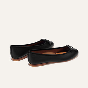 The Demi - Black Nappa by margaux-development: handmade in Spain, these black leather ballet flats feature brown soles and delicate front bows, showcased on a simple light background.
