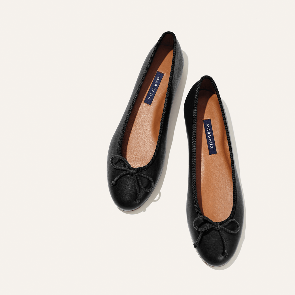 A pair of The Demi - Black Nappa ballet flats by margaux-development, handmade in Spain with tan insoles labeled MARGAUX and small bows on the toes, shown side by side on a light background.