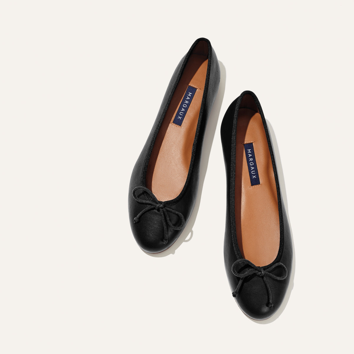 A pair of The Demi - Black Nappa ballet flats by margaux-development, handmade in Spain with tan insoles labeled MARGAUX and small bows on the toes, shown side by side on a light background.