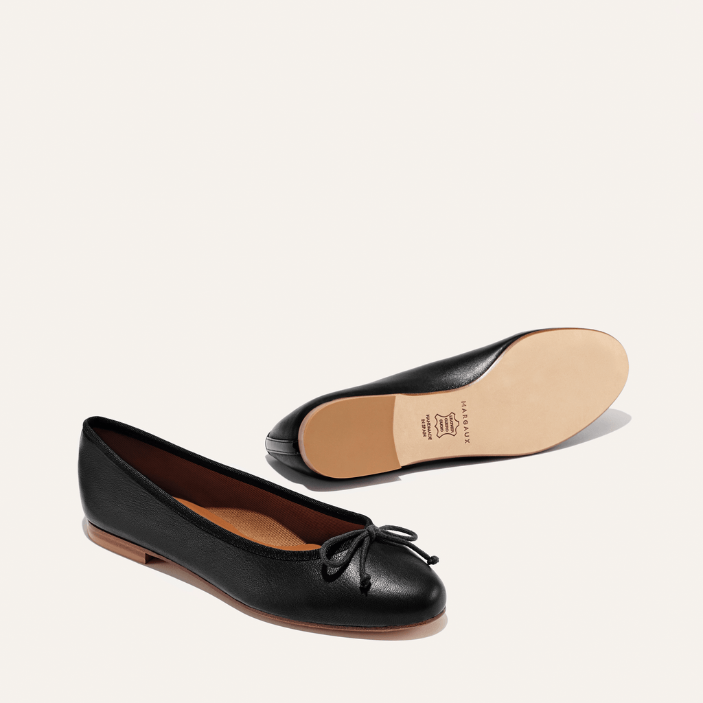 A pair of The Demi - Black Nappa ballet flats by margaux-development, handmade in Spain from black leather with a small bow on the toe; one shoe stands upright, the other on its side showing the tan sole.
