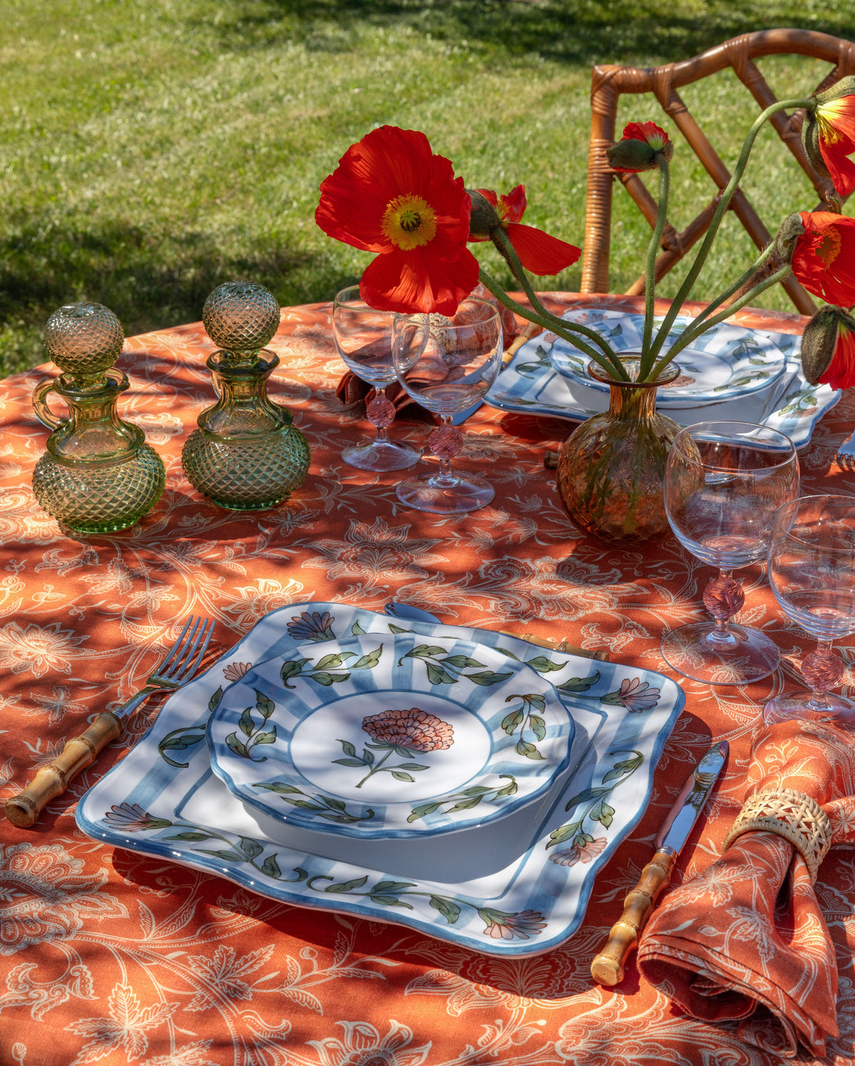 Orange floral tablecloth covers an outdoor table set with glassware and floral plates beside red flowers
