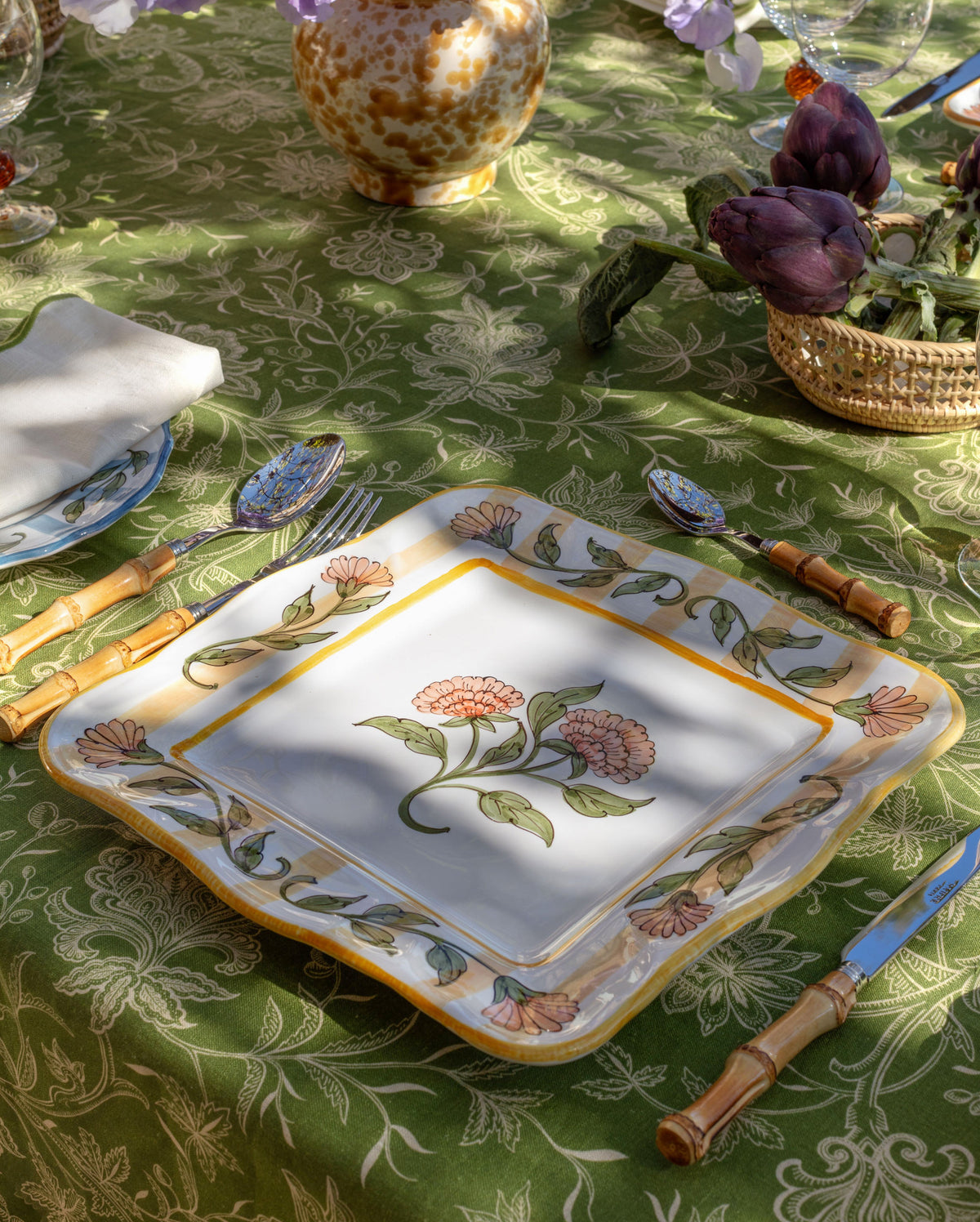 Tablescape featuring a square floral plate on a patterned green cloth with flowers, glassware, and bamboo handled utensils