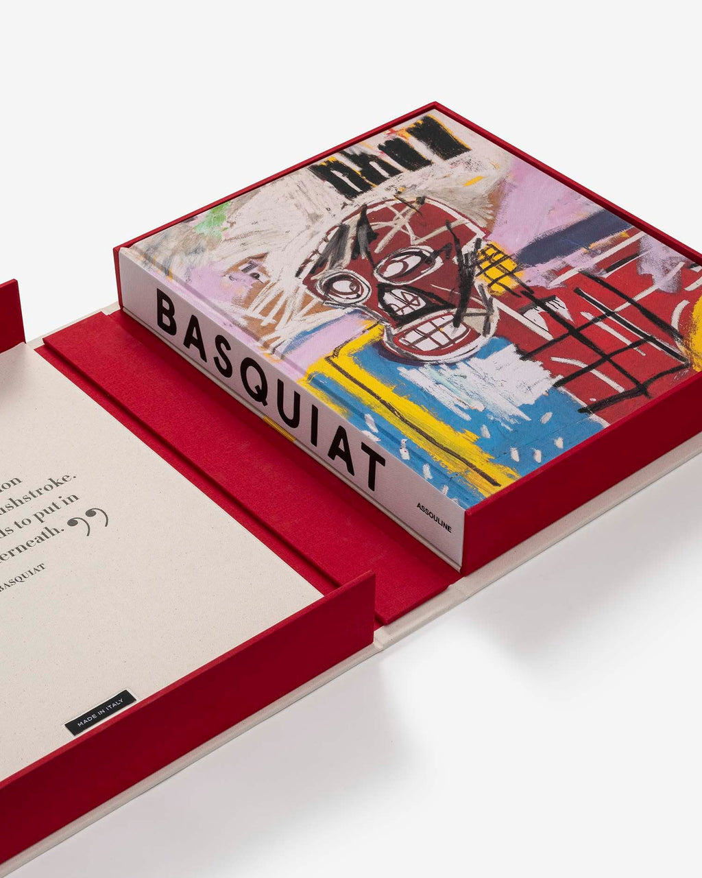 Basquiat: The World of Jean-Michel by Assouline: book in the open red clamshell, angled overhead view; the book cover reproduces a vivid Basquiat painting — a yellow and pink skull-like face against an abstract field of red, blue, and yellow with the word pneu inscribed in black — with BASQUIAT on the white spine; the clamshell's cream interior lid bears an italic Basquiat quote; a Made in Italy label is visible inside the box
