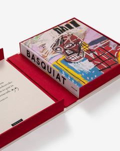 Basquiat: The World of Jean-Michel by Assouline: book in the open red clamshell, angled overhead view; the book cover reproduces a vivid Basquiat painting — a yellow and pink skull-like face against an abstract field of red, blue, and yellow with the word pneu inscribed in black — with BASQUIAT on the white spine; the clamshell's cream interior lid bears an italic Basquiat quote; a Made in Italy label is visible inside the box