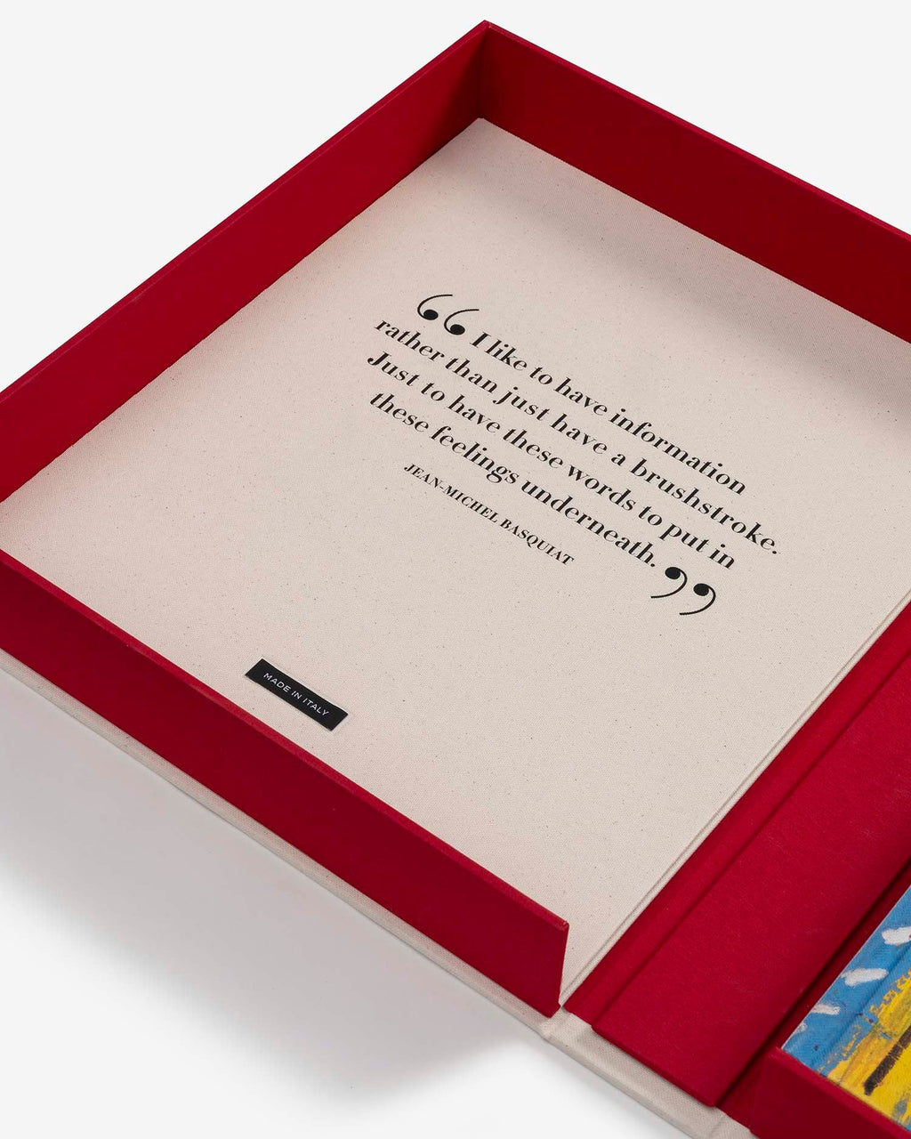 Basquiat: The World of Jean-Michel by Assouline: close-up overhead view of the open red clamshell interior lid; the cream panel bears a Basquiat quote in black italic serif type: I like to have information rather than just have a brushstroke. Just to have these words to put in these feelings underneath. — Jean-Michel Basquiat; a small Made in Italy label is centered at the base; a corner of the colorful book cover is visible at lower right