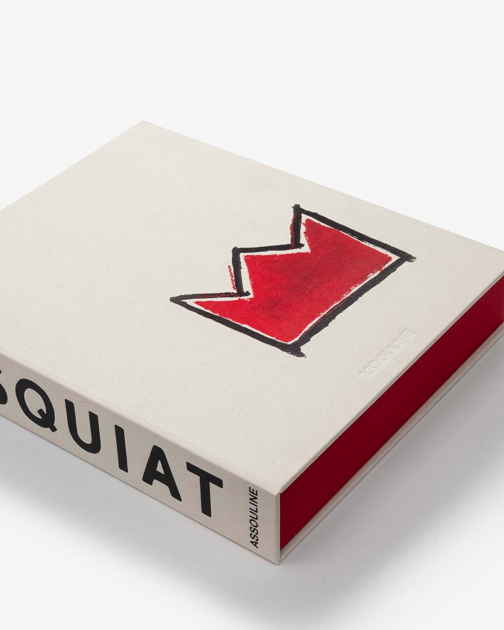 Basquiat: The World of Jean-Michel by Assouline: close-up angled overhead view of the book resting on the open red slipcase; the cream canvas cover shows the red crown motif; BASQUIAT and ASSOULINE on the spine; the red slipcase trim frames the right and bottom edges