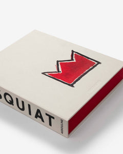 Basquiat: The World of Jean-Michel by Assouline: close-up angled overhead view of the book resting on the open red slipcase; the cream canvas cover shows the red crown motif; BASQUIAT and ASSOULINE on the spine; the red slipcase trim frames the right and bottom edges