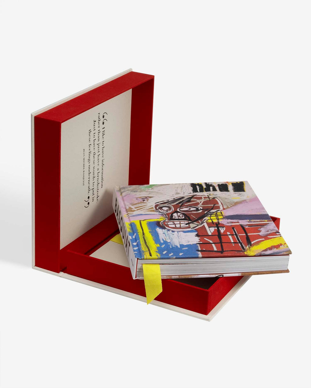 Basquiat: The World of Jean-Michel by Assouline: full product presentation — the open cream-and-red clamshell stands vertically in the background displaying the Basquiat quote on its ivory lid; in the foreground the book rests in the red tray, its colorful cover showing a Basquiat skull painting in vivid yellow, red, and blue with the word pneu; a yellow grosgrain ribbon bookmark trails from the binding